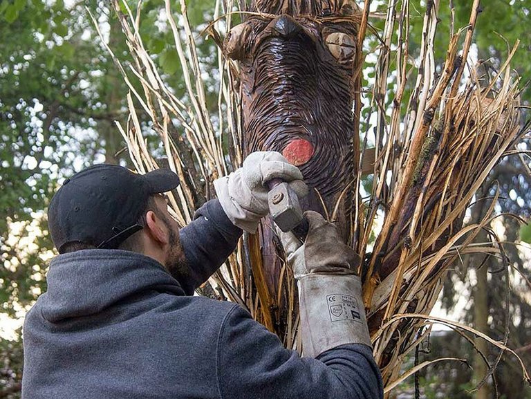 Ein Mann meißelt mit Arbeitshandschuhen (aber ohne Schutzbrille) an einer Skulptur aus Holz